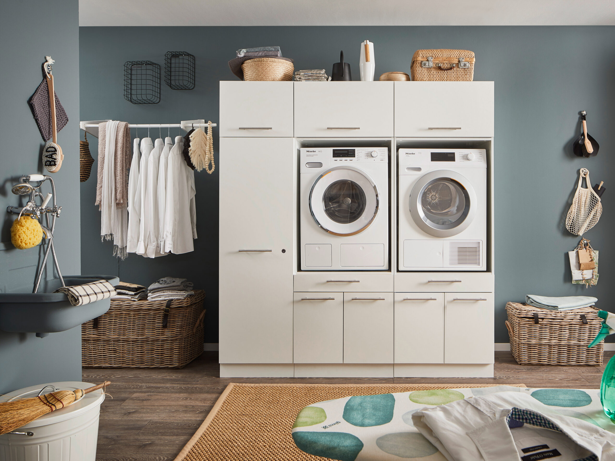 Ein Waschraum mit Waschmaschine und Trockner im weißen Möbelmodul, Kleiderstange mit Hemden, Körben und einem Wandwaschtrog.