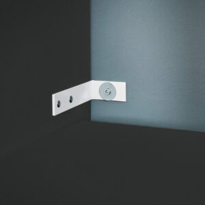 Ein weißes Metallscharnier mit drei Schrauben ist an einer dunklen Oberfläche und einer hellen Wand montiert.