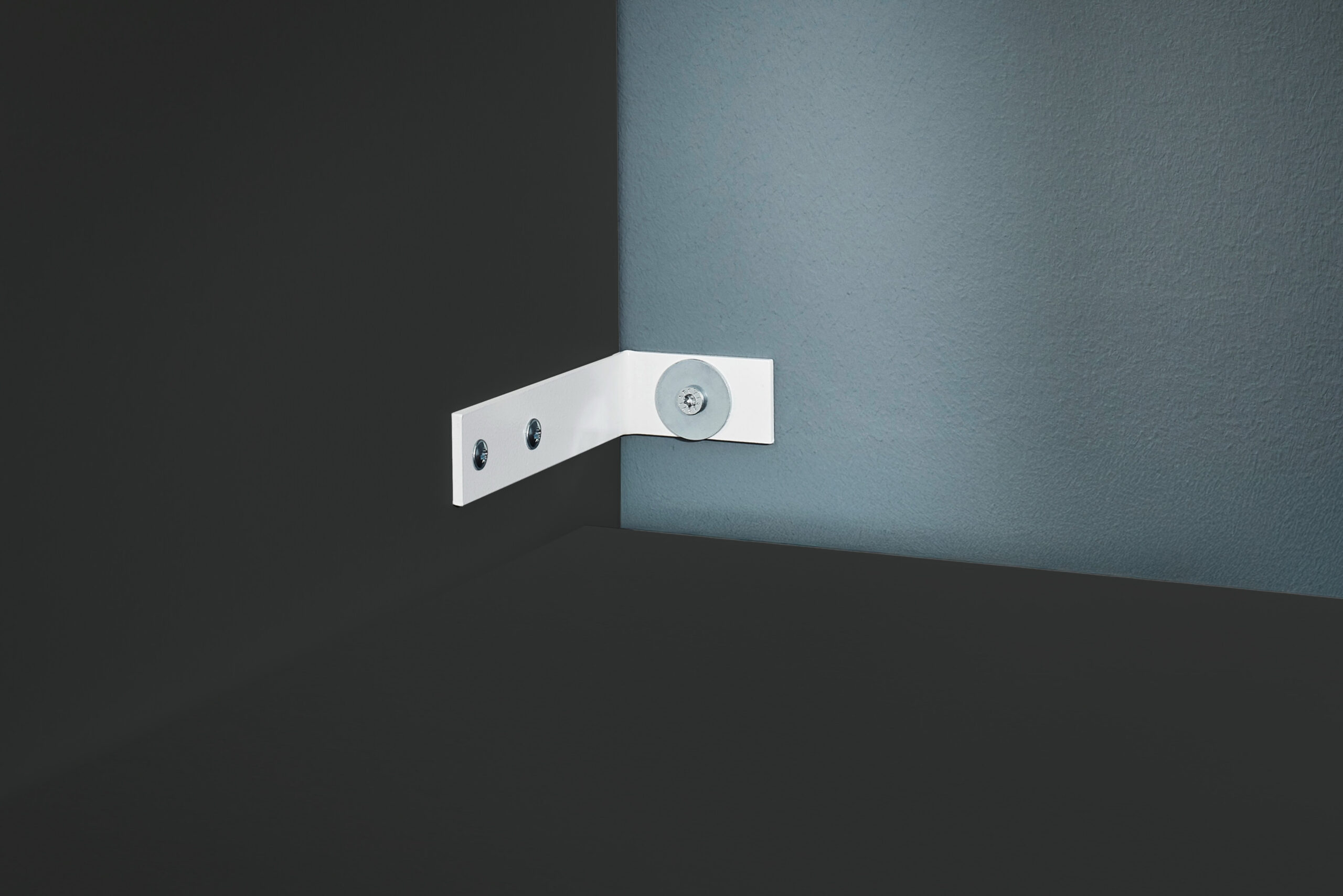 Ein weißes Metallscharnier mit drei Schrauben ist an einer dunklen Oberfläche und einer hellen Wand montiert.