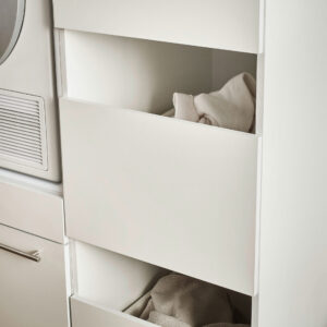 Ein weißer Schrank mit offenen Fächern, in denen helle Textilien liegen, steht neben einem Trockner in einem modernen Hauswirtschaftsraum.
