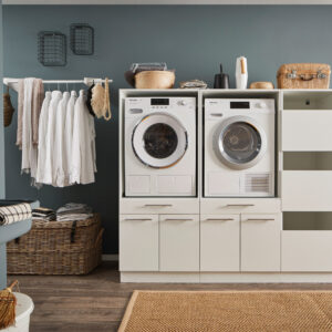 Ein Waschraum mit Waschmaschine und Trockner im weißen Möbelmodul, offenen Regalfächern, Kleiderstange mit weißen Hemden, Körben und einem Wandwaschtrog.