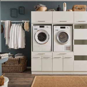 Ein Waschraum mit Waschmaschine und Trockner im weißen Möbelmodul, offenen Stauraumfächern, Kleiderstange mit weißen Hemden, Körben und einem Wandwaschtrog.