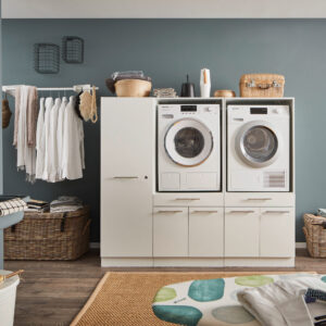 Ein Waschraum mit Waschmaschine und Trockner im weißen Möbelmodul, Kleiderstange mit weißen Hemden, Körben und einem Wandwaschtrog.