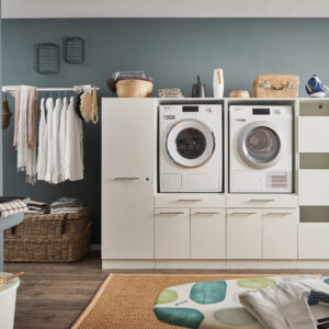 Ein Waschraum mit Waschmaschine und Trockner im weißen Möbelmodul, Kleiderstange mit Hemden, offenen Regalfächern, Körben und einem Wandwaschtrog.