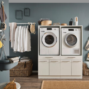 Ein Waschraum mit Waschmaschine und Trockner in einem weißen Möbelmodul, flankiert von Körben, einer Kleiderstange und einem Waschtrog an der Wand.