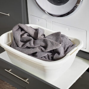 Eine weiße Wanne mit grauen Textilien steht auf einer herausgezogenen Ablage unter einer Waschmaschine in einem modern eingerichteten Waschbereich.