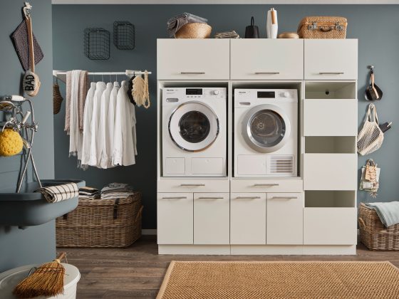 Ein Waschraum mit Waschmaschine und Trockner im wei&szlig;en M&ouml;belmodul, offenen Stauraumf&auml;chern, Kleiderstange mit wei&szlig;en Hemden, K&ouml;rben und einem Wandwaschtrog.