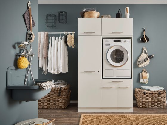 Ein Waschraum mit eingebauter Waschmaschine in einem weißen Möbelmodul, Kleiderstange mit weißen Hemden, Körben und einem Wandwaschtrog.